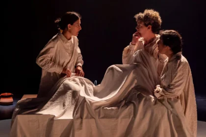 Tres actrius amb camisa de dormir al voltant d'un llit en una escena de ‘Rogles de cançons i misèria’, de Cactus Teatre