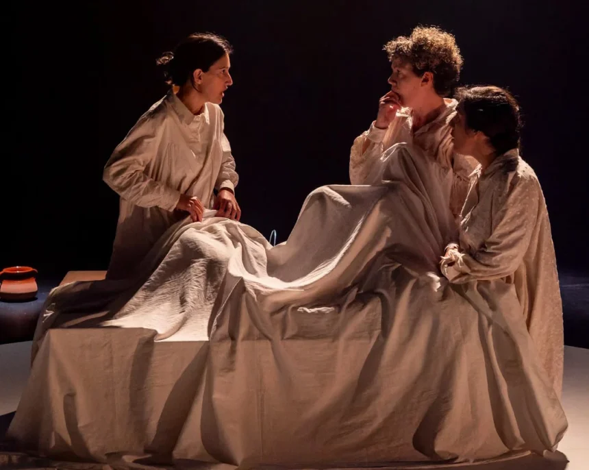 Tres actrius amb camisa de dormir al voltant d'un llit en una escena de ‘Rogles de cançons i misèria’, de Cactus Teatre