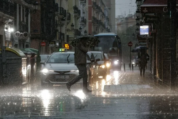 Un peatón con paraguas cruza una calle bajo una fuerte lluvia mientras circulan coches con las luces encendidas