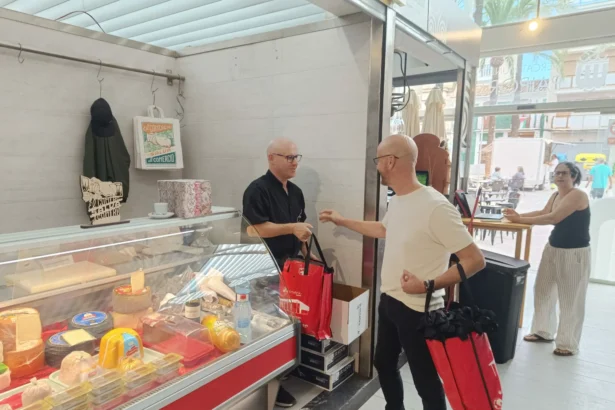 Dos hombres junto a un puesto de mercado con quesos y embutidos entregan bolsas rojas a un cliente