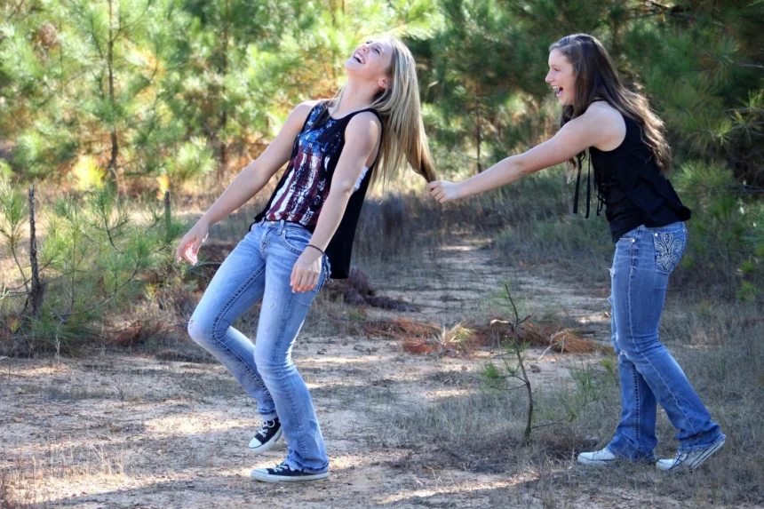 Dos adolescentes ríe a carcajadas mientras una tira del pelo a la otra en un camino rodeado de pinos
