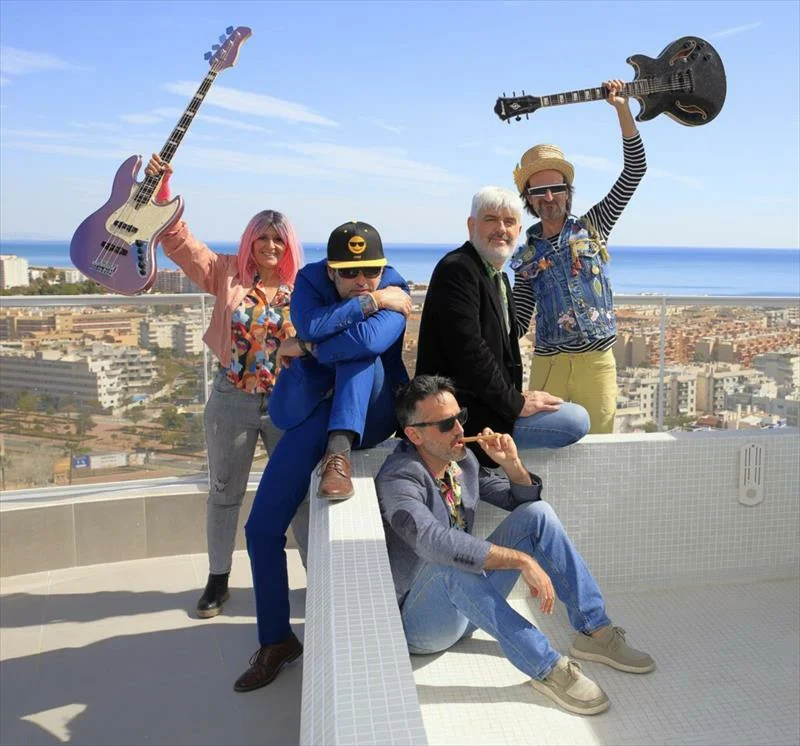 Cinco músicos posan en una azotea con una guitarra y un bajo en alto, con el mar al fondo