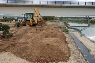Retroexcavadora trabajando en la orilla de un cauce junto a un puente en Sueca