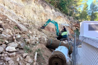 Excavadora trabajando en el talud rocoso junto al depósito de agua potable de Sumacàrcer, con rollos de malla de protección
